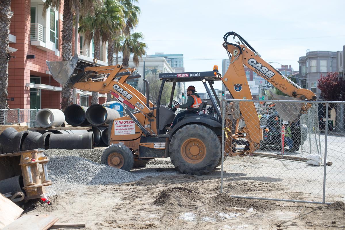 Construction worker operating backhoe to backfill utility trench on urban street with palm trees and buildings visible.