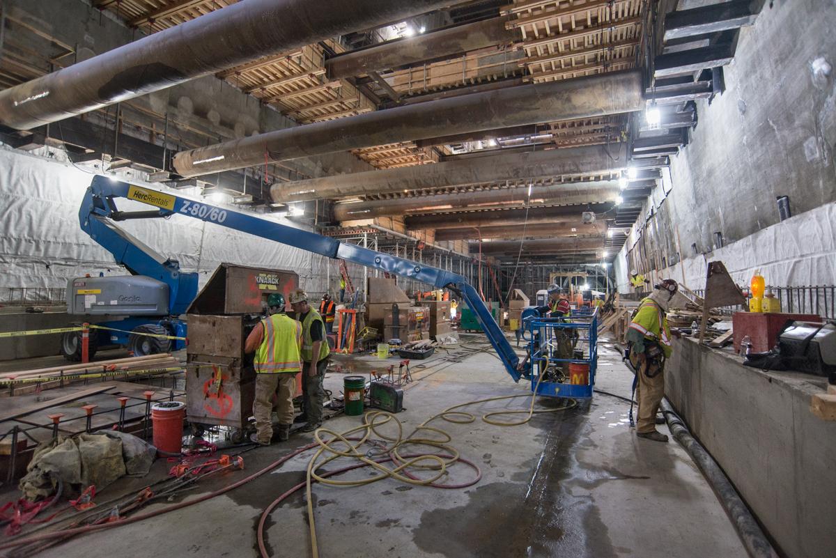 Workers in safety vests install waterproofing and bracing inside an underground transit station tunnel.