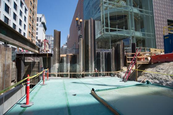 Construction site on 4th Street with protective barriers, equipment, and concrete work area surrounded by downtown buildings.