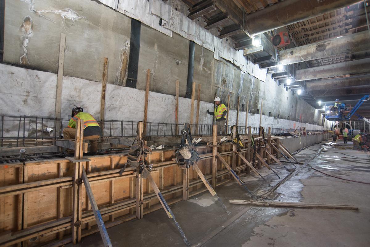 Workers construct rebar cages and concrete forms inside a large underground transit station box.