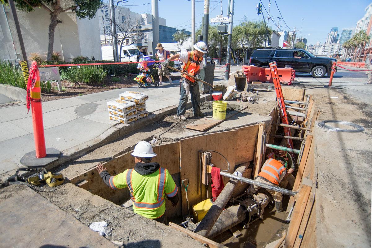 Construction workers in safety vests perform utility work in an excavated street trench on a city street.