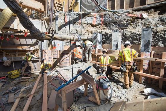 Construction workers in safety vests build wooden shoring and formwork at an urban construction site.