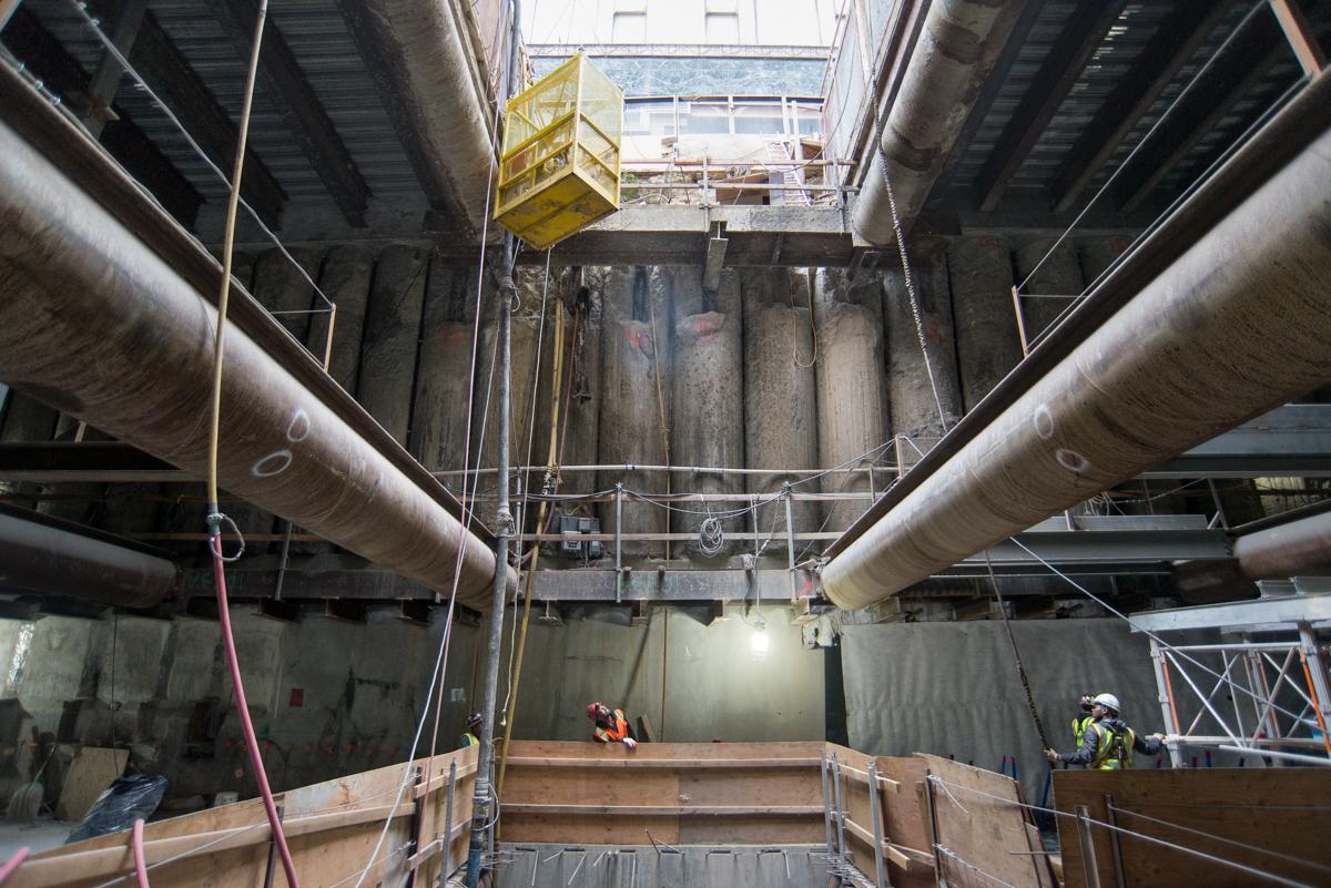 Construction workers on scaffolding inside underground Central Subway tunnel shaft with large pipes and concrete walls.