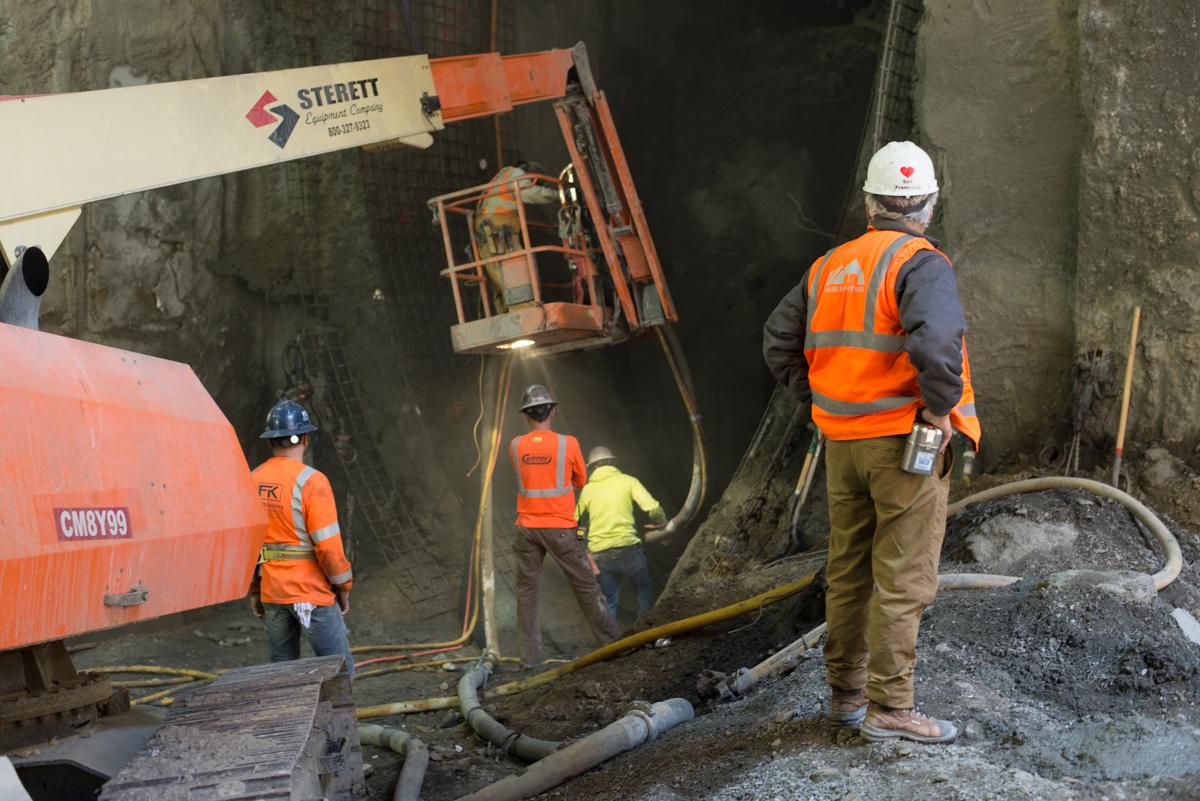 Construction workers in safety vests operate a concrete pump truck at an underground transit station construction site.