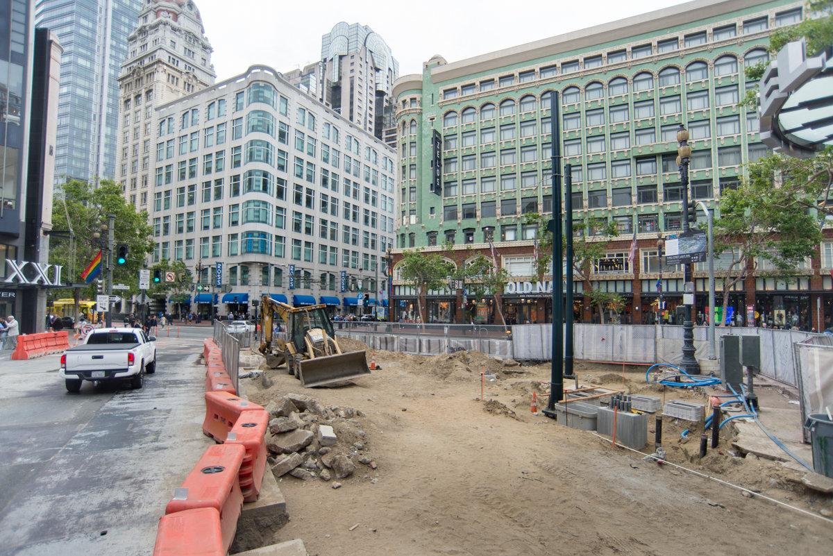 Construction site at urban intersection with excavation equipment, barriers, and historic buildings in background