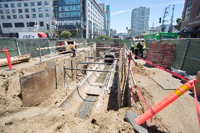 Construction site showing excavated trench with sewer line installation between buildings in urban area.
