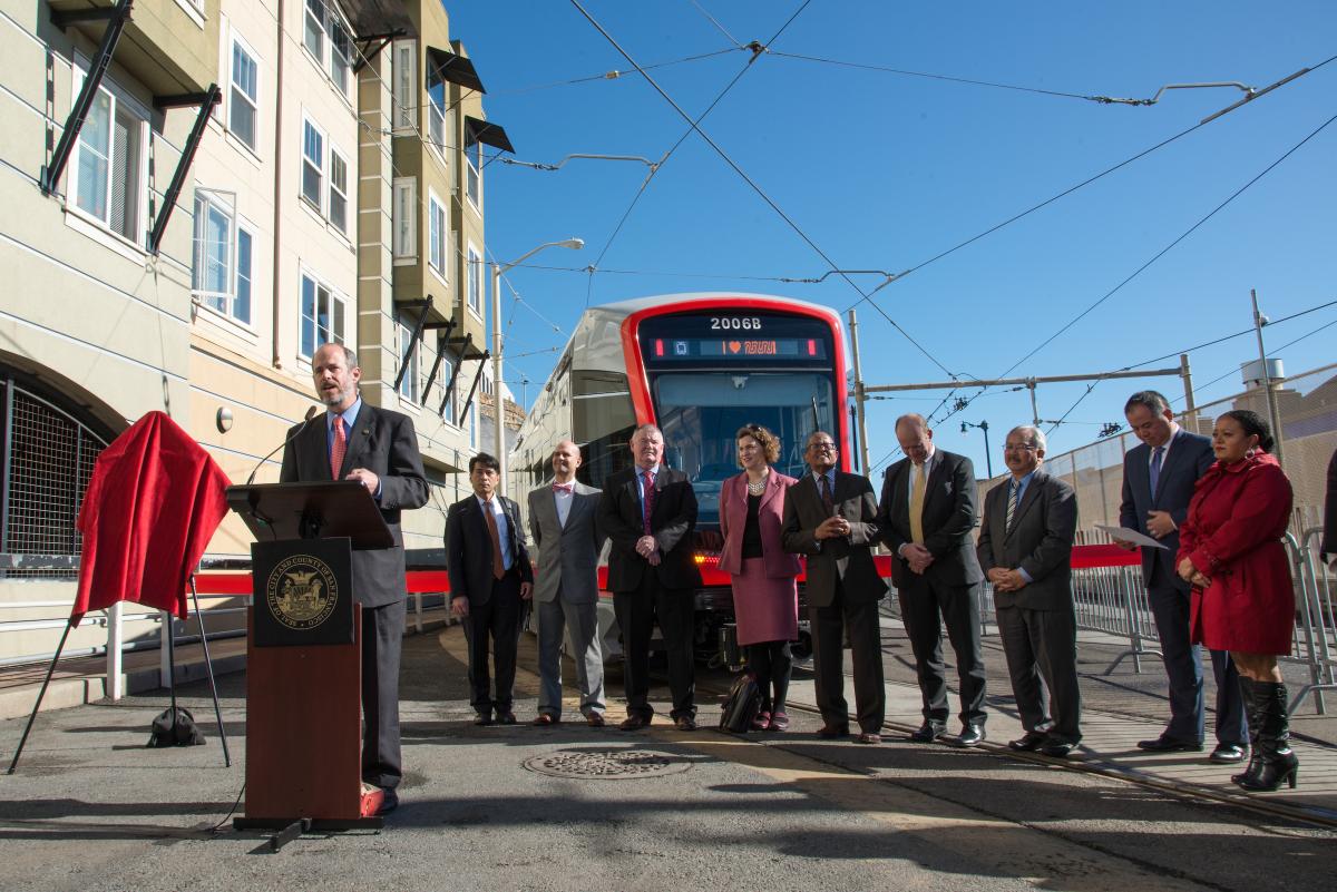 Official ceremony with dignitaries at podium beside new red Muni light rail train on city street.