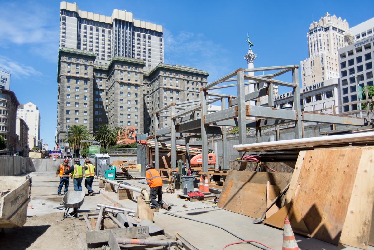 Construction workers and equipment at downtown transit station excavation site with steel bracing and concrete work underway.