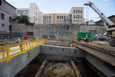 Construction excavation site with concrete foundation, yellow safety barriers, crane, and surrounding buildings on Stockton Street.