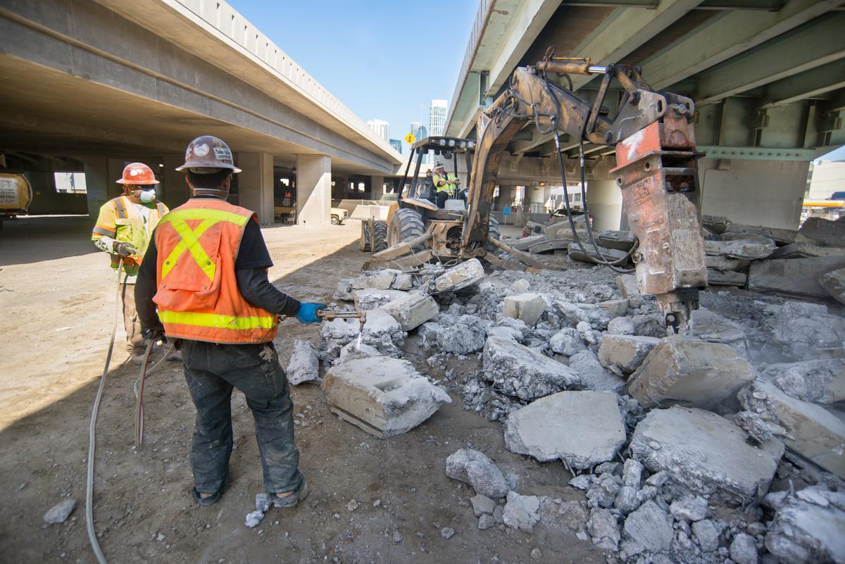 Workers in safety gear use jackhammer and torch to demolish concrete tunnel sections under highway overpass.