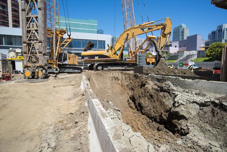 Yellow excavator operating at construction site with concrete foundation wall and earthen pit for material storage.