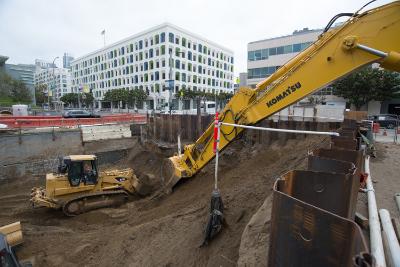 Excavator removing soil from trench at Yerba Buena/Moscone Station construction site with city buildings in background.