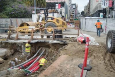 Construction workers install underground utilities in excavated street trench with heavy equipment nearby.