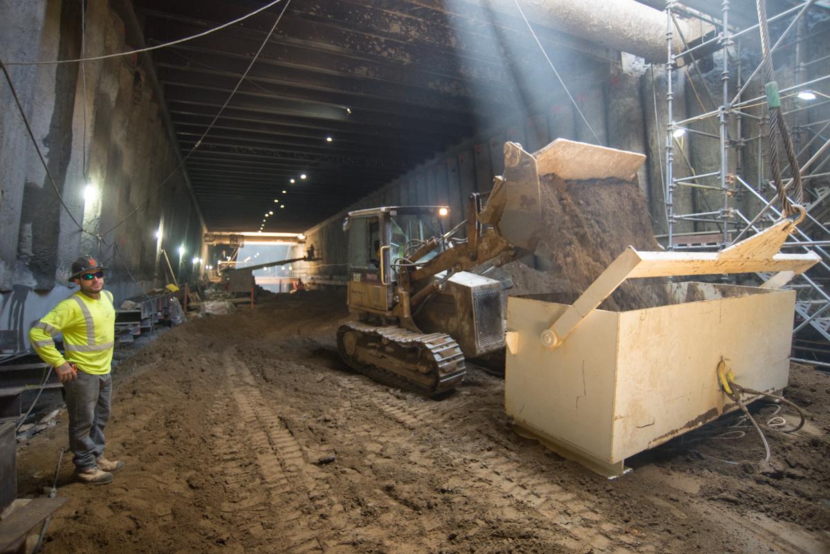 Construction worker in yellow vest observing excavator loading soil into steel bucket inside underground transit station.