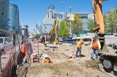 Construction site with excavation equipment, workers in safety vests, and urban buildings in background.