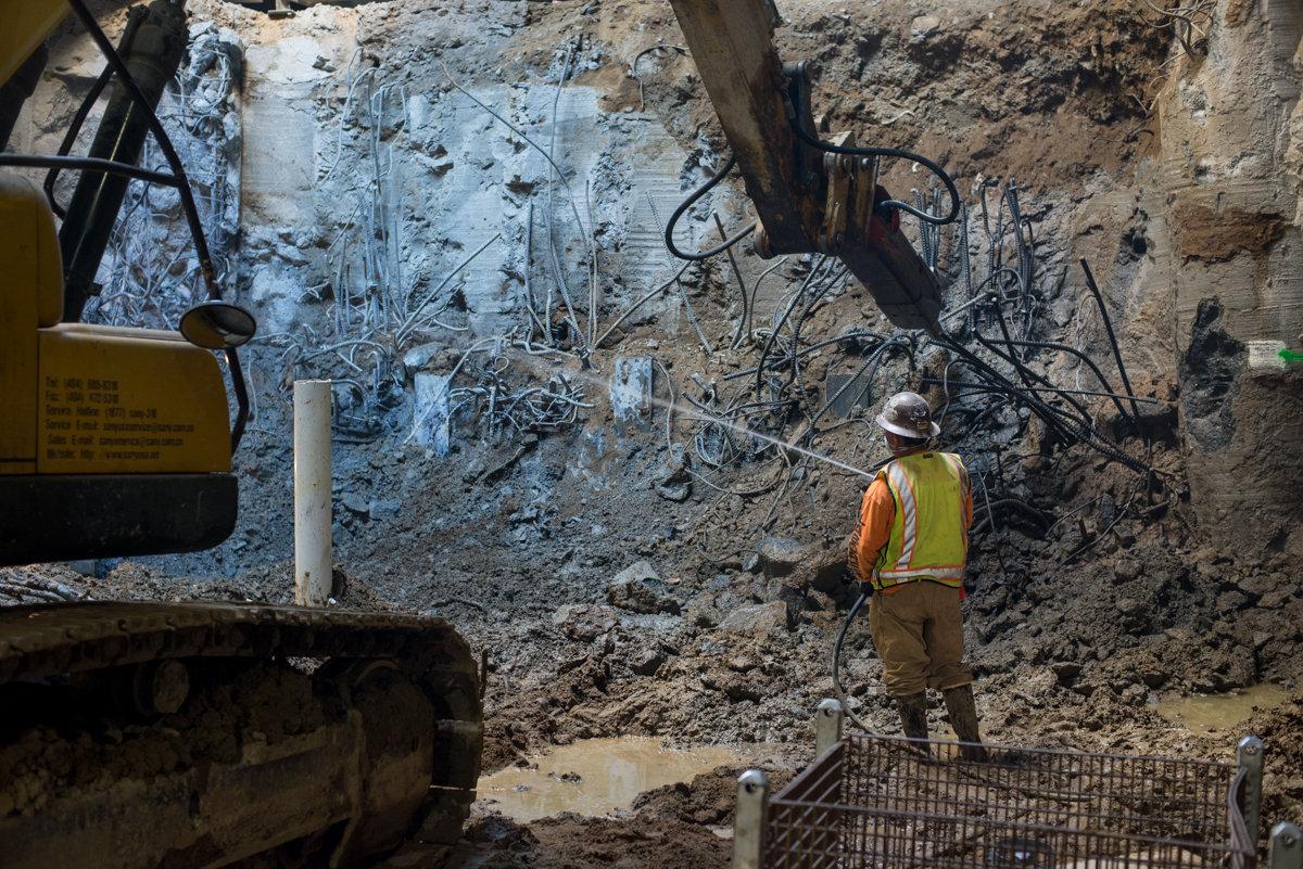 Construction worker in safety gear observing excavation site with heavy equipment and temporary steel bracing on rock wall.