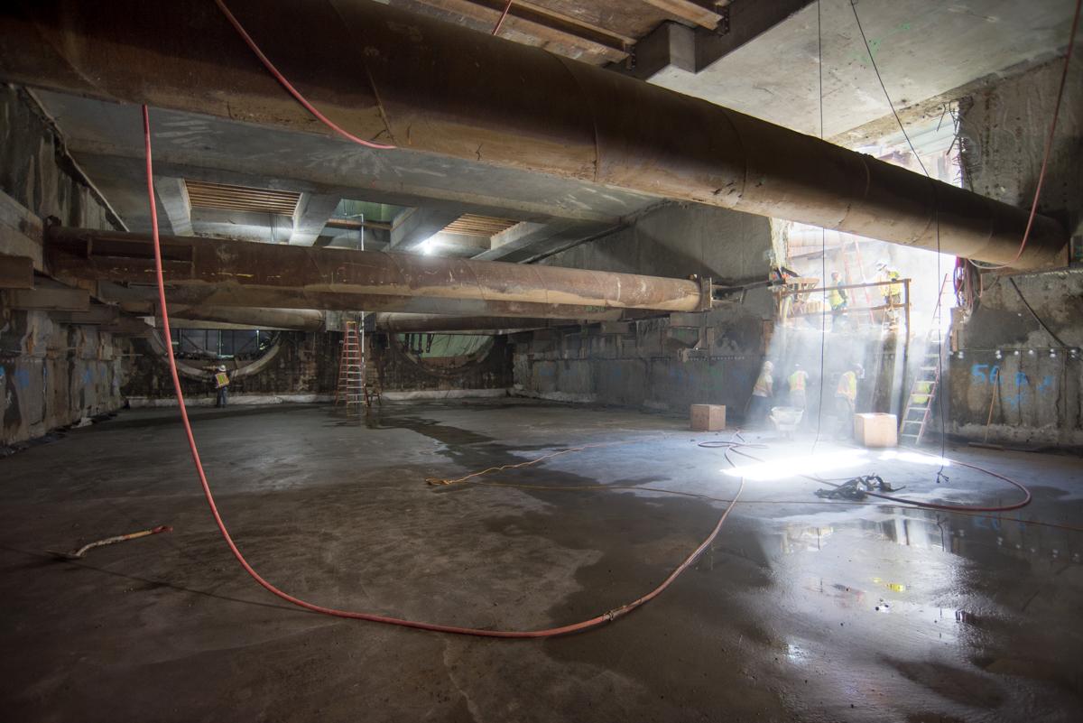 Interior concrete construction space under elevated highway with red hose and work lights.