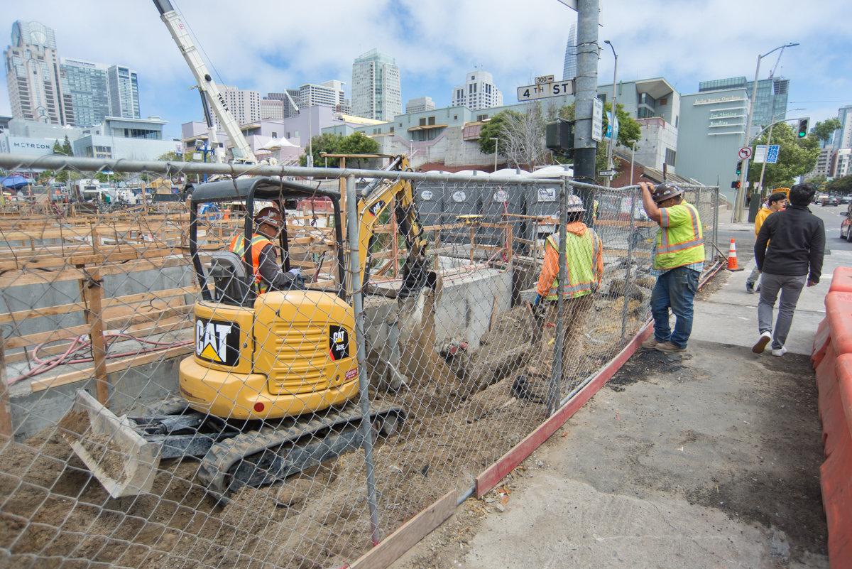 Construction workers remove formwork and bracing from Yerba Buena/Moscone Station headhouse with downtown skyline visible.