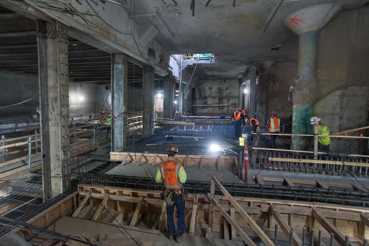 Construction workers in safety vests work on structural steel and concrete formwork inside an underground transit station excavation.