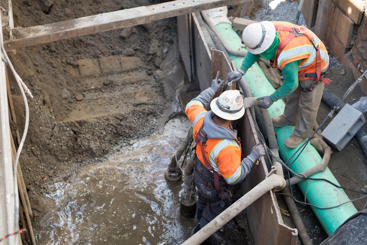 Two workers in hard hats and safety vests install green pipes in an underground excavation trench with water and soil visible.