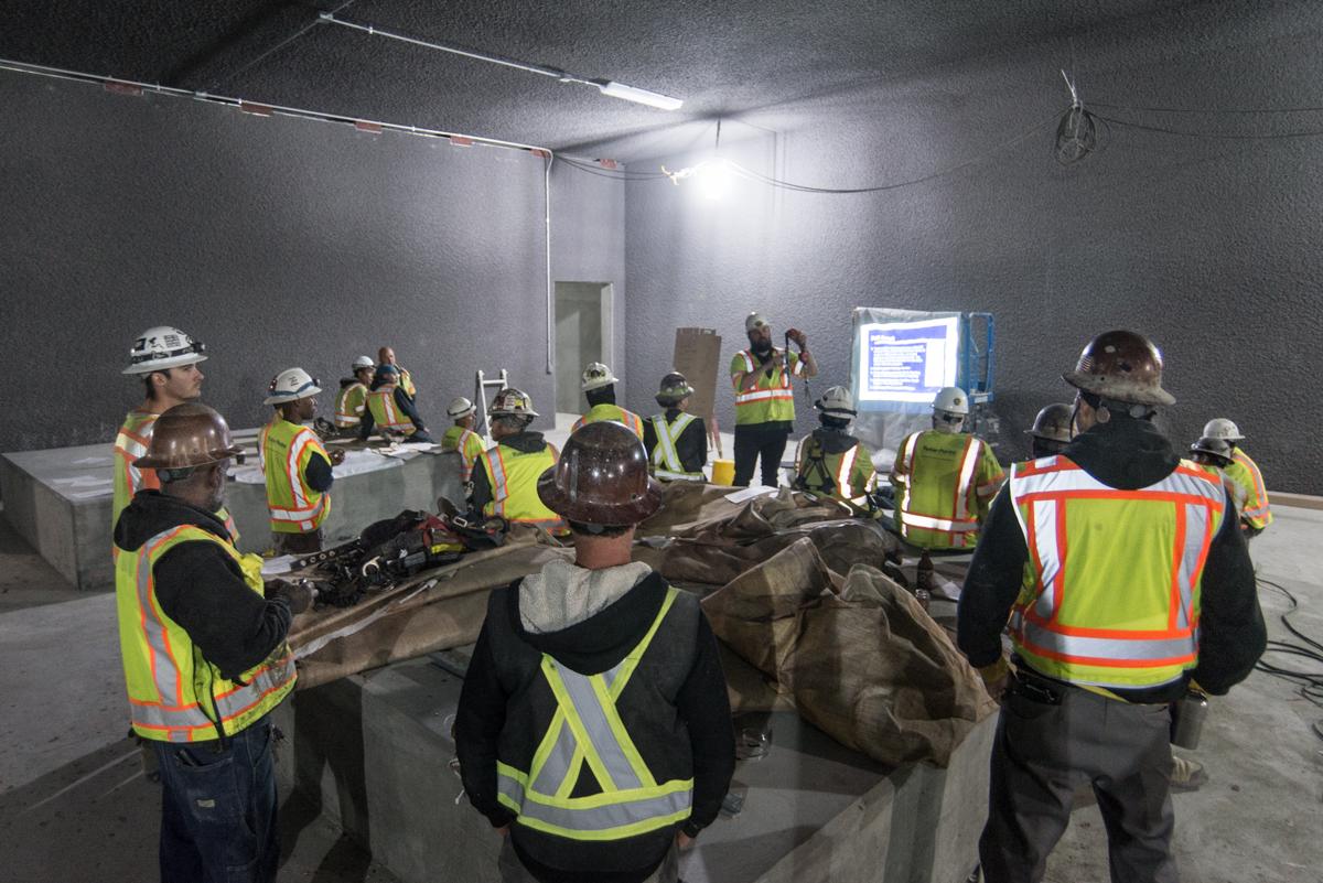 Construction workers in safety vests and hard hats gather around rebar cages during a fall safety training session in an underground transit station.