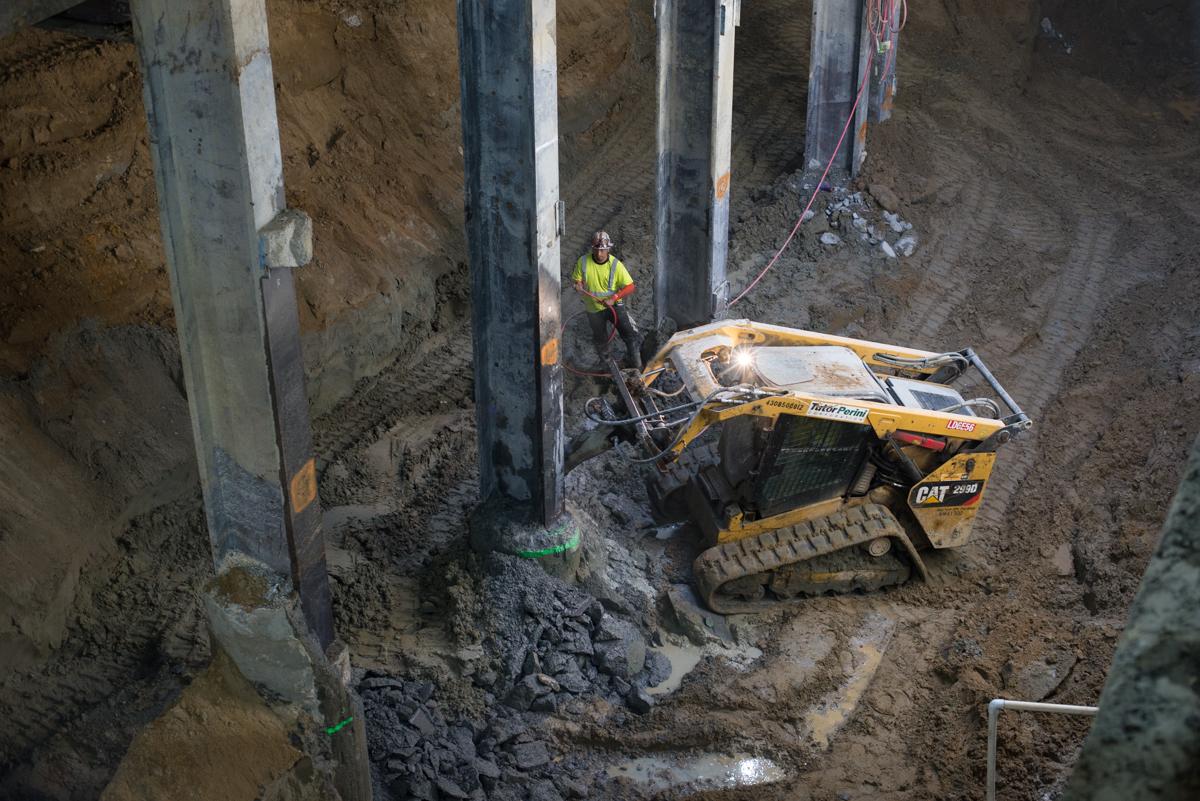 Bobcat operator removes concrete around vertical support beam in underground transit construction site.