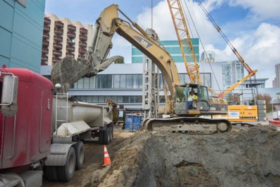 Excavator loading dirt and debris onto truck at urban construction site with buildings and cranes in background.