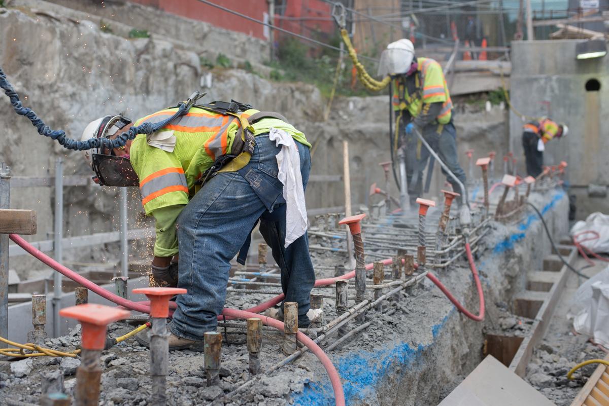 Construction worker in safety gear operating pneumatic tool on concrete foundation wall at transit construction site.