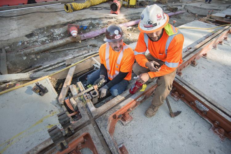 Two construction workers in orange safety vests and hard hats prepare welding equipment on railroad tracks at a transit work site.