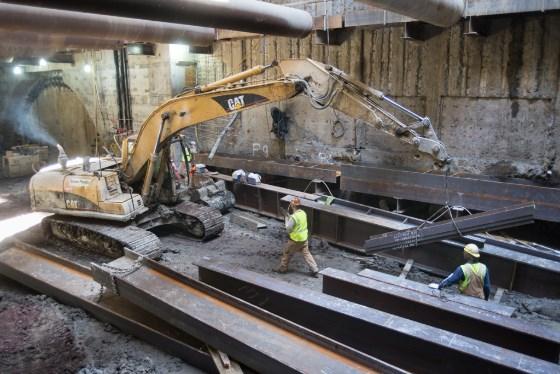 CAT excavator loads steel beams into truck bed during underground station construction work.