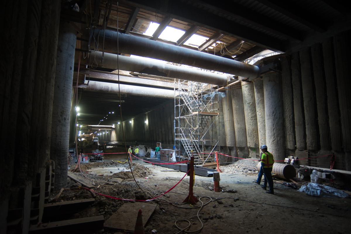 Underground transit station under construction with concrete columns, temporary bracing, and workers in safety gear.