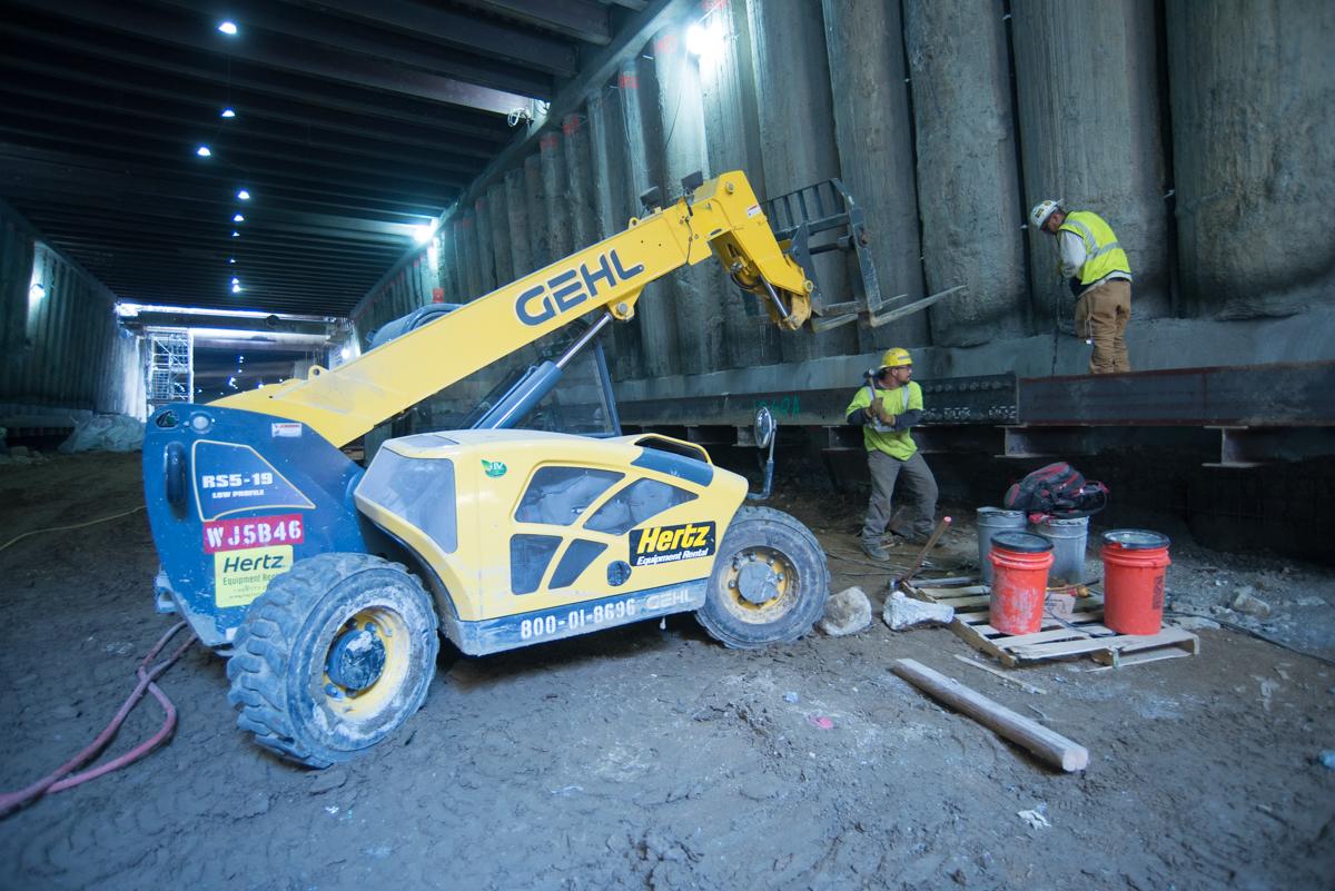 Telehandler lifting steel beam while workers guide placement in underground station construction area.