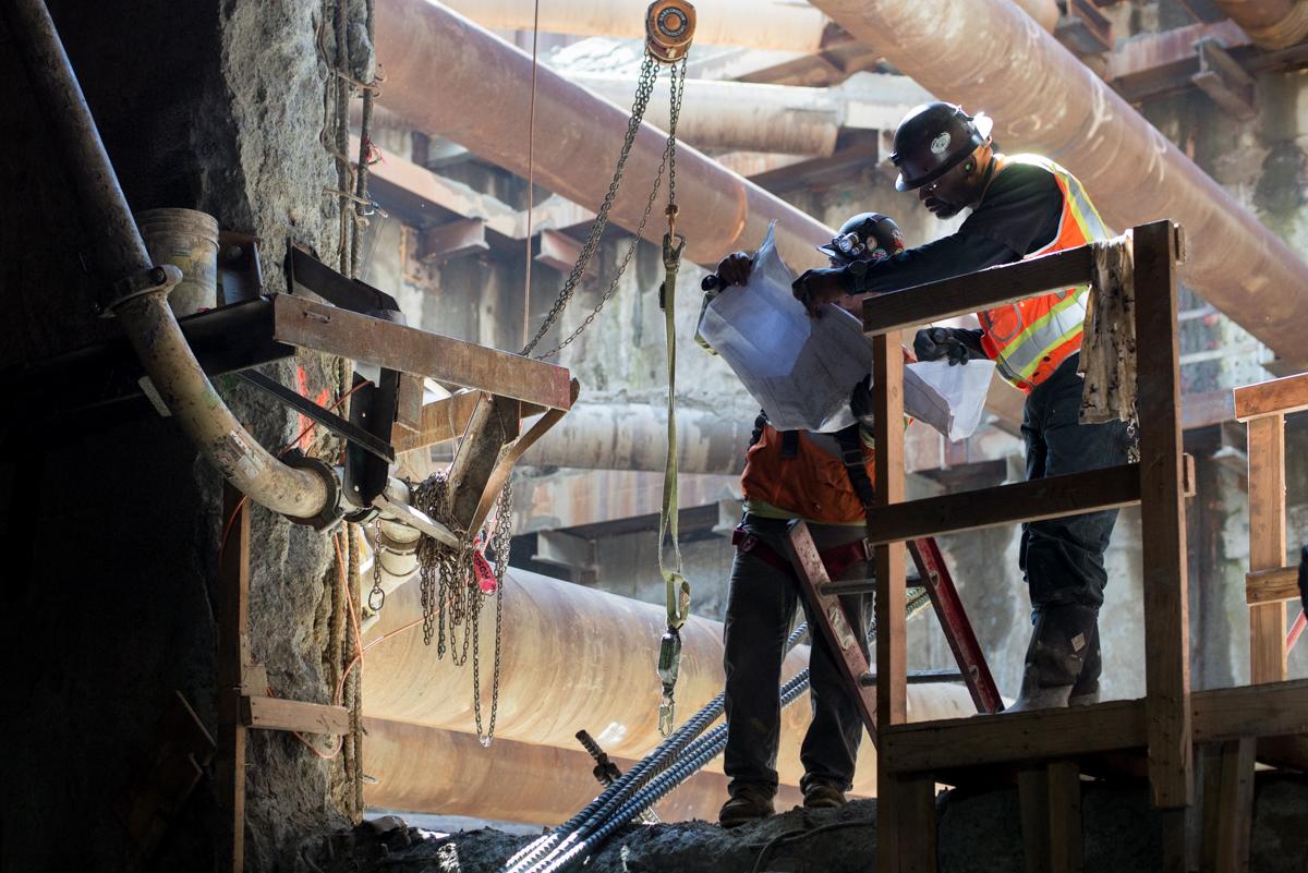 Workers in safety gear review construction plans while building rebar cage at station platform level.