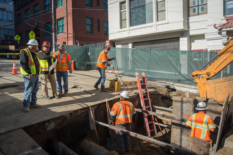 Construction workers in safety vests inspect underground utility work on an urban street with excavation equipment and protective barriers.