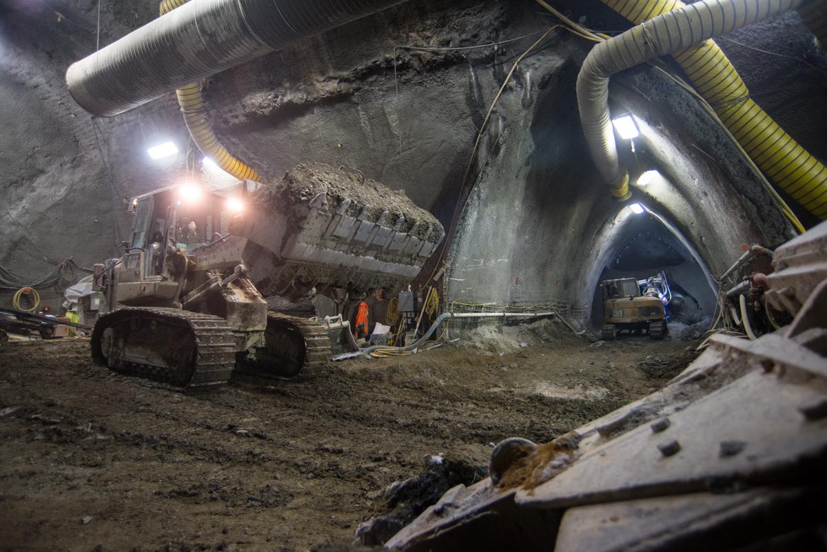 Bulldozer moving excavated material inside underground station cavern with ventilation ducts and work lights.
