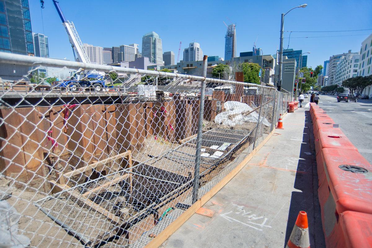 Construction site with chain-link fencing along urban street, city skyline visible in background.
