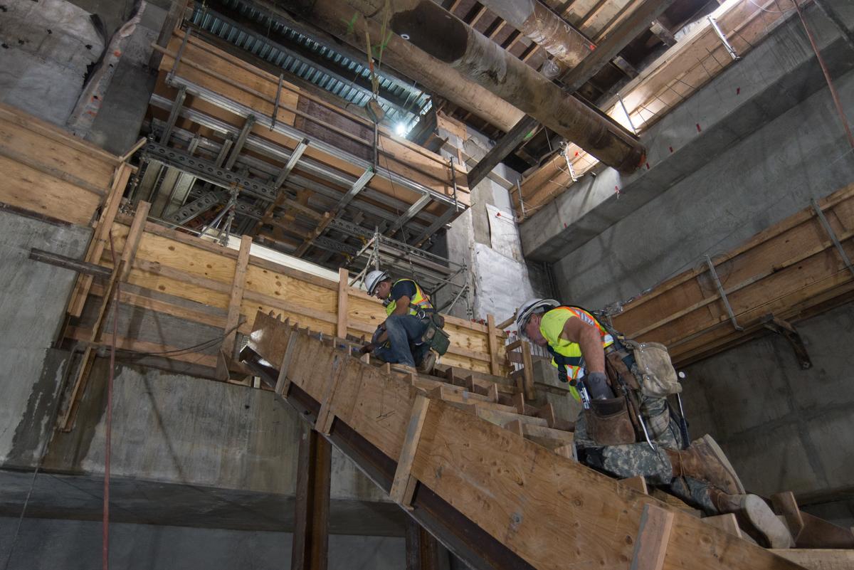 Two construction workers in safety gear install wooden formwork and steel framing for an internal staircase in a transit station.