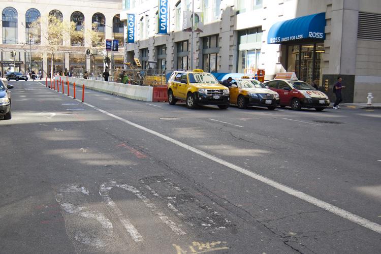 Temporary taxi stand on 4th Street with yellow cabs lined up behind red and white barriers, urban street with buildings and storefronts.
