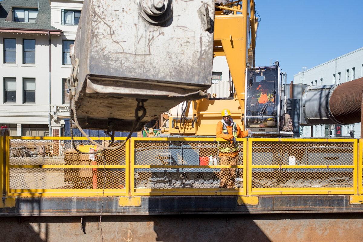 Heavy construction crane lowers large concrete barrel vault section onto yellow platform at urban transit construction site.