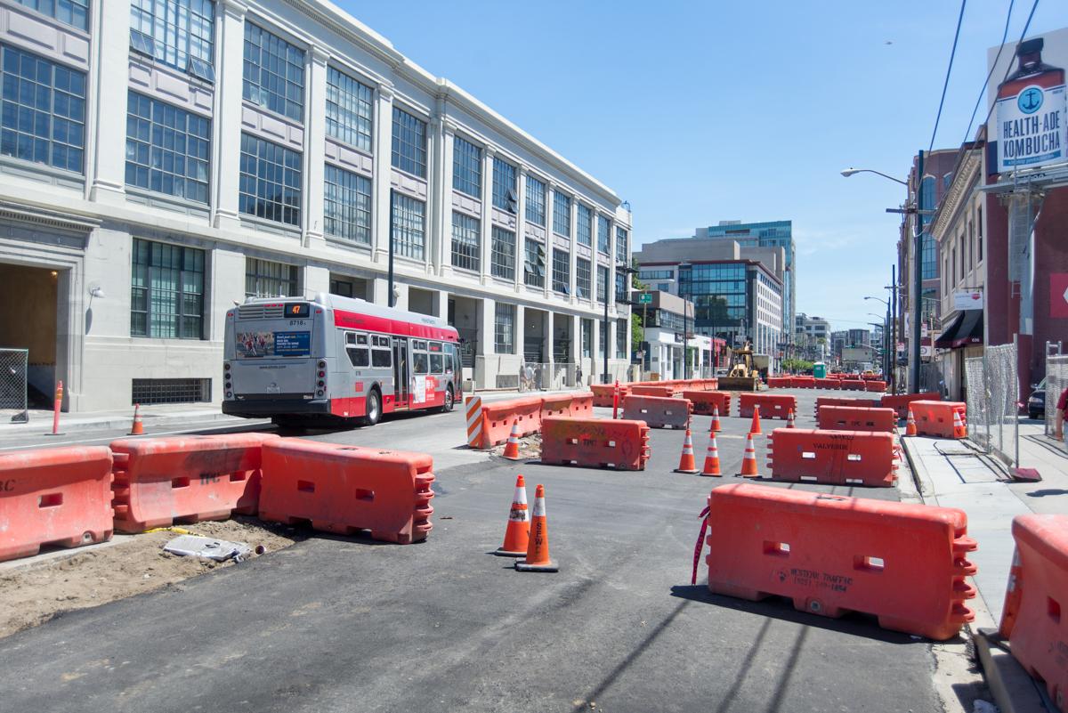 Construction work zone on 4th Street with red barriers, traffic cones, and transit bus passing by.