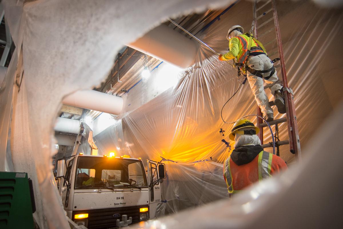 Worker suspended on ropes paints interior structural supports inside transit station while truck waits below.
