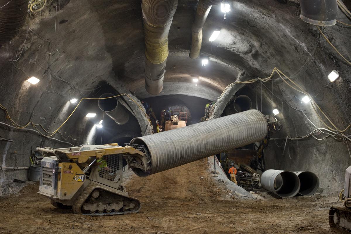 Tracked loader installing large spiral ventilation duct in underground transit tunnel cavern with LED work lights.