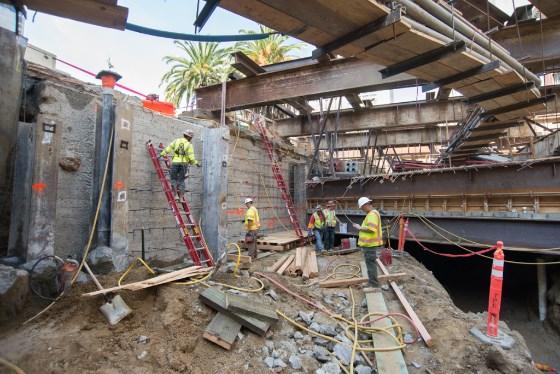 Construction workers in safety gear excavating street infrastructure with exposed utilities and equipment at urban intersection.