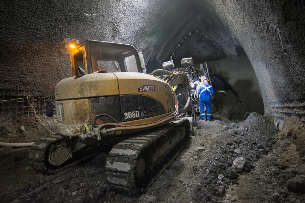 Excavator operating inside underground tunnel cavern with workers in blue safety gear nearby.