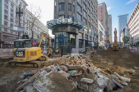 Construction excavation at urban intersection with yellow CAT equipment and exposed rock debris, surrounded by multi-story buildings.