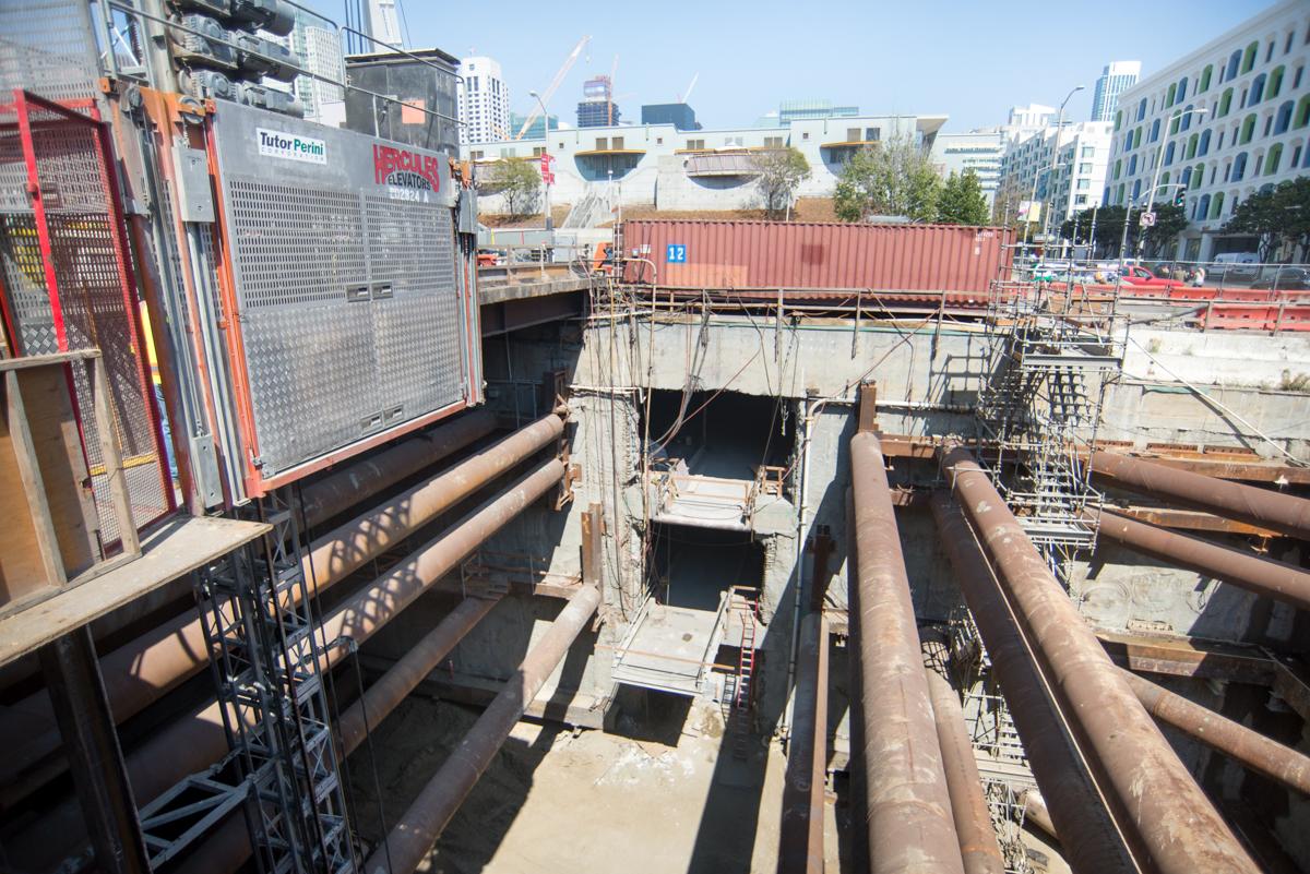 Construction site showing excavated station box with large pipes and steel reinforcement under 4th Street.