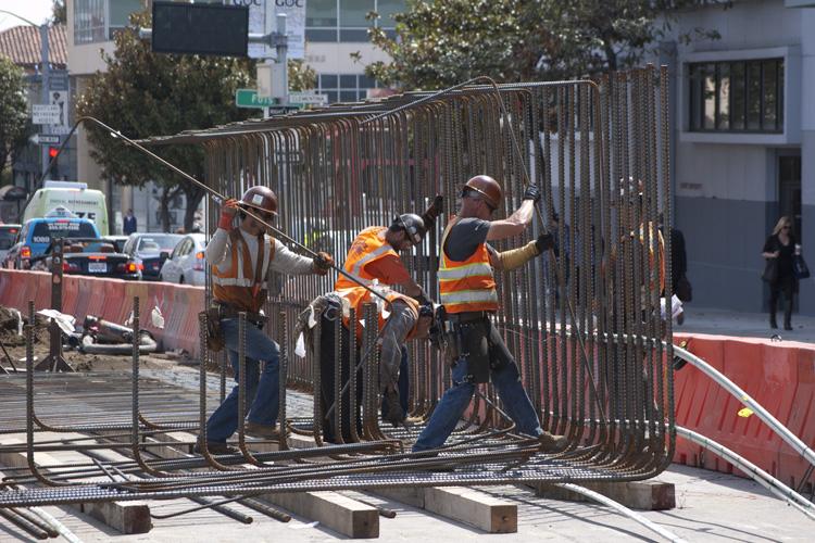Construction workers in safety vests and hard hats position a large steel rebar cage on a city street surrounded by barriers.