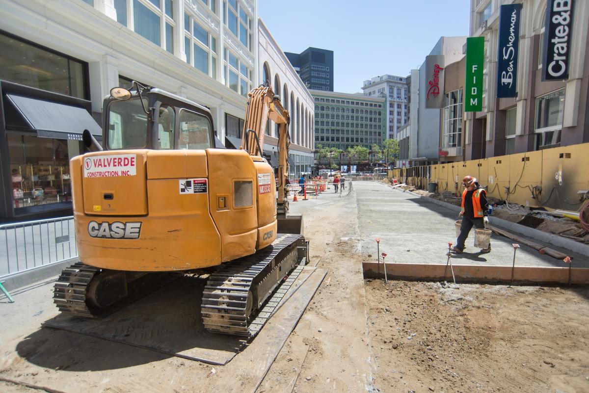 Construction site on Stockton Street with orange excavator and workers, downtown buildings visible.