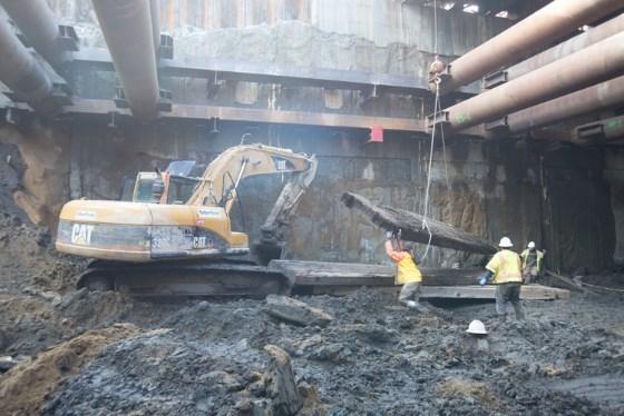 Excavator lowering wooden support beams into underground transit station headhouse during construction.