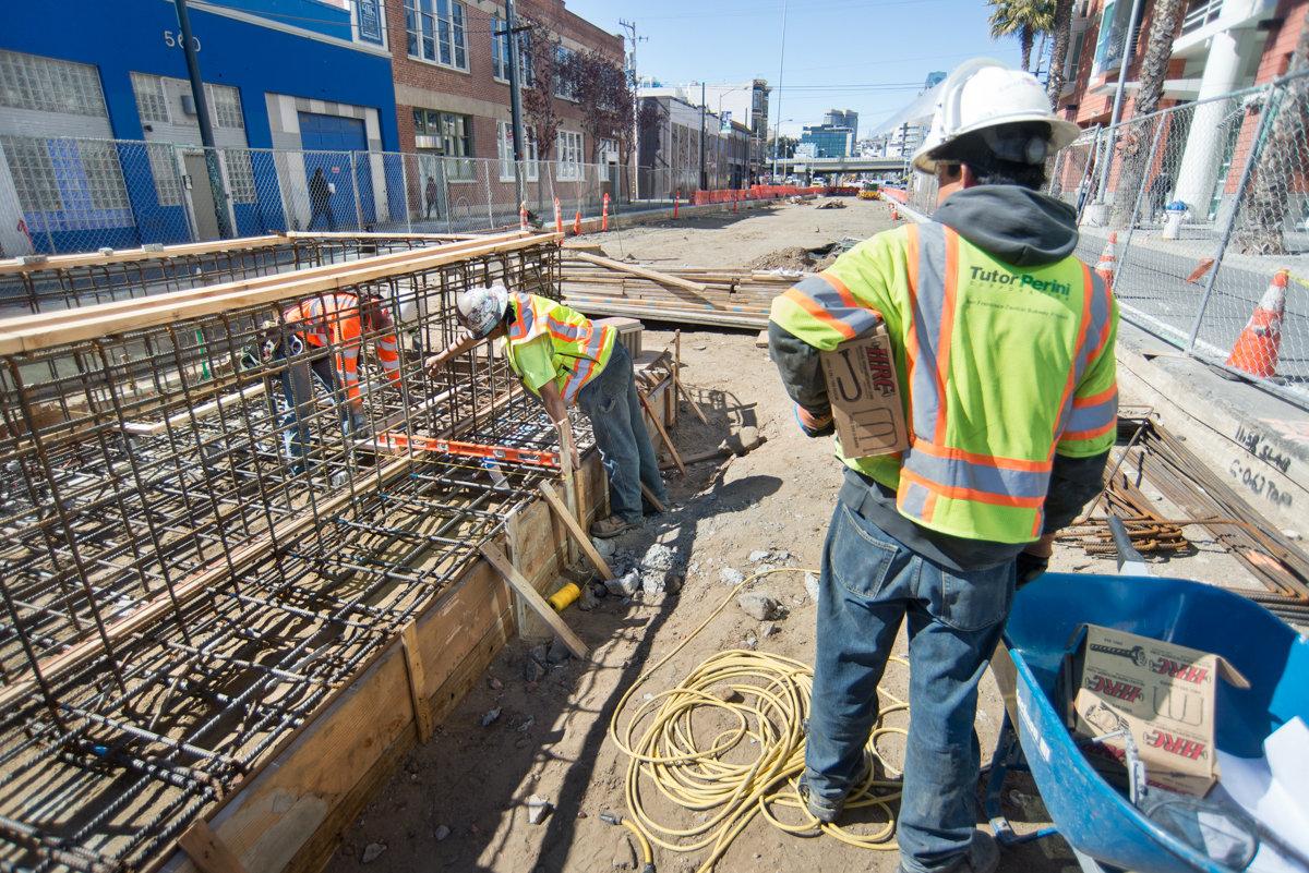 Construction workers install rebar cages in concrete formwork at transit tunnel station site.